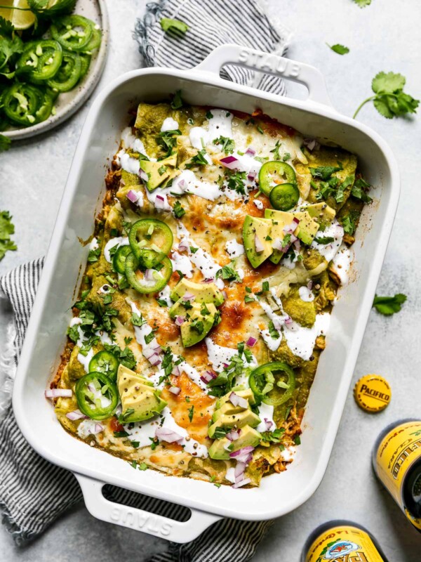 Baked green enchiladas covered in cheeese, red onion, avocado, & sliced jalapeno. The baking dish sits atop a striped gray linen napkin on a light blue surface, surrounded by cilantro leaves, Pacifico beer, & a small plate of garnishes (lime wedges, cilantro, sliced jalapeno, etc.)
