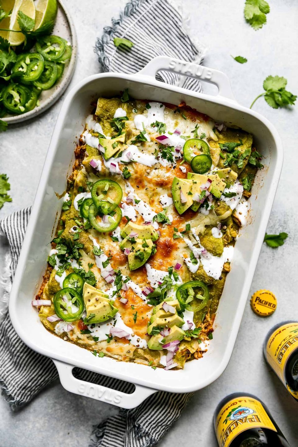 Baked green enchiladas covered in cheeese, red onion, avocado, & sliced jalapeno. The baking dish sits atop a striped gray linen napkin on a light blue surface, surrounded by cilantro leaves, Pacifico beer, & a small plate of garnishes (lime wedges, cilantro, sliced jalapeno, etc.)