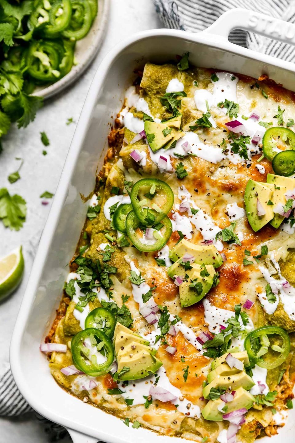 Baked green enchiladas covered in cheeese, red onion, avocado, & sliced jalapeno. The baking dish sits atop a striped gray linen napkin on a light blue surface, surrounded by cilantro leaves, & a small plate of garnishes (lime wedges, cilantro, sliced jalapeno, etc.)