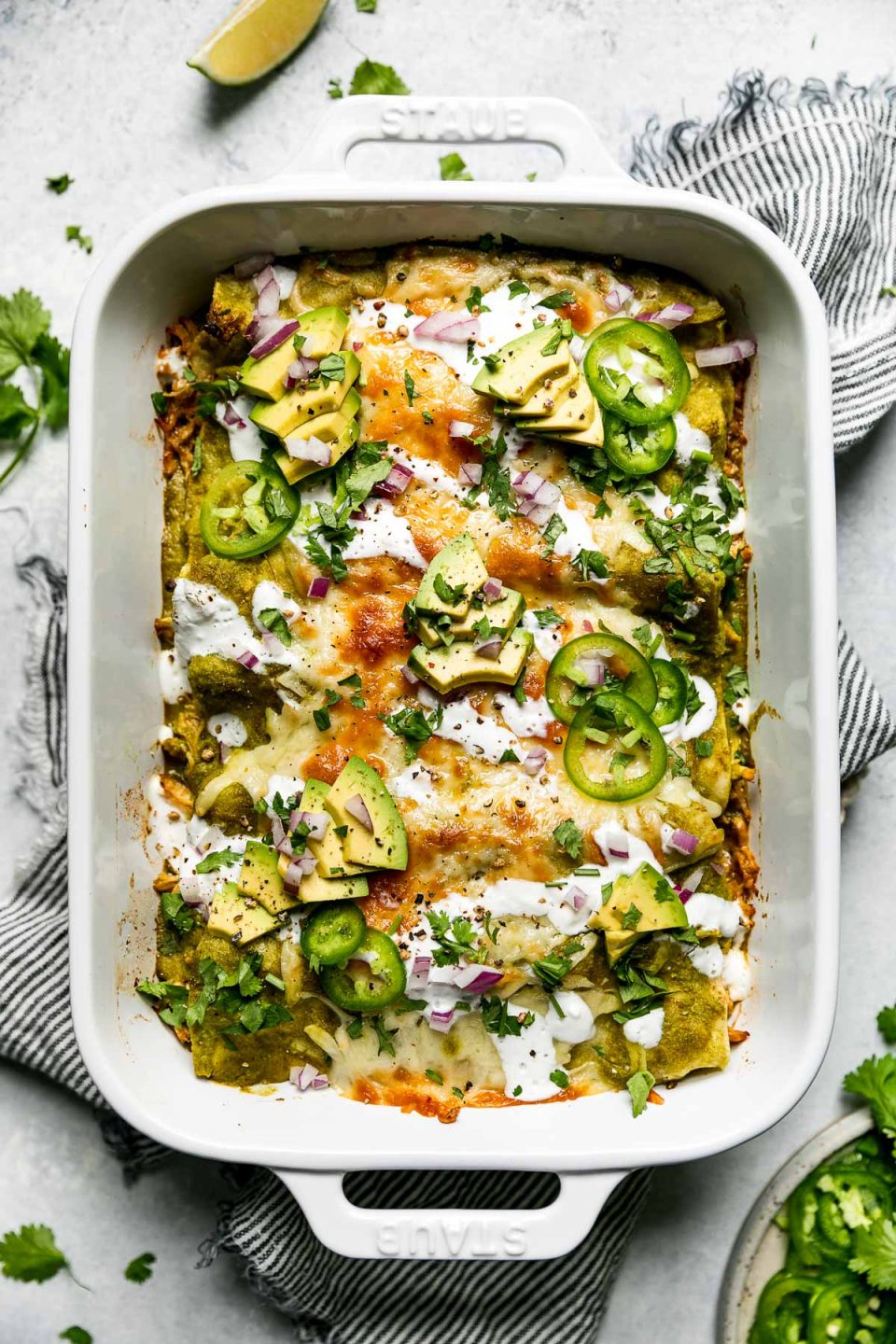 Baked green enchiladas covered in cheeese, red onion, avocado, & sliced jalapeno. The baking dish sits atop a striped gray linen napkin on a light blue surface, surrounded by cilantro leaves, lime wedges, & a small plate of garnishes (lime wedges, cilantro, sliced jalapeno, etc.)