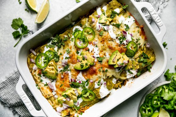 Baked green enchiladas covered in cheeese, red onion, avocado, & sliced jalapeno. The baking dish sits atop a striped gray linen napkin on a light blue surface, surrounded by cilantro leaves, lime wedges, & a small plate of garnishes (lime wedges, cilantro, sliced jalapeno, etc.)