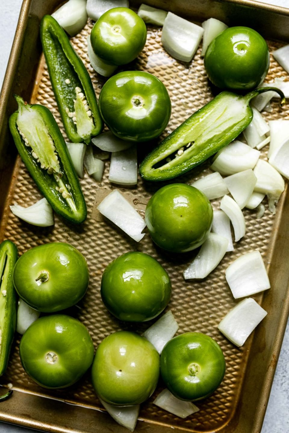 Homemade green enchilada sauce ingredients on a small sheet pan atop a light blue surface before roasting – tomatillos, halved peppers, & chopped onion.