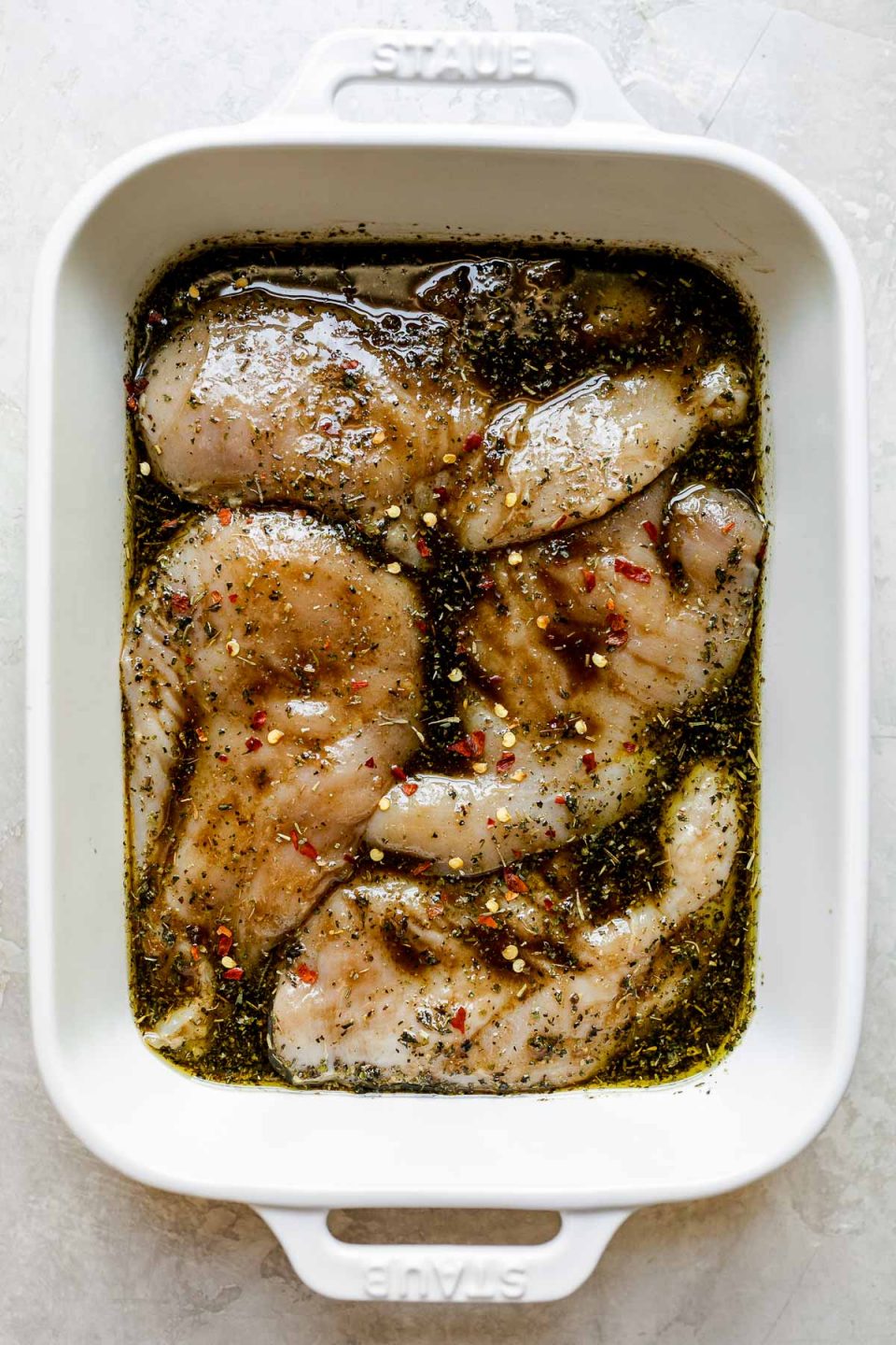 Boneless, skinless chicken breasts rest in a white Staub baking dish filled with simple balsamic marinade. The baking sits on top of a white textured surface.