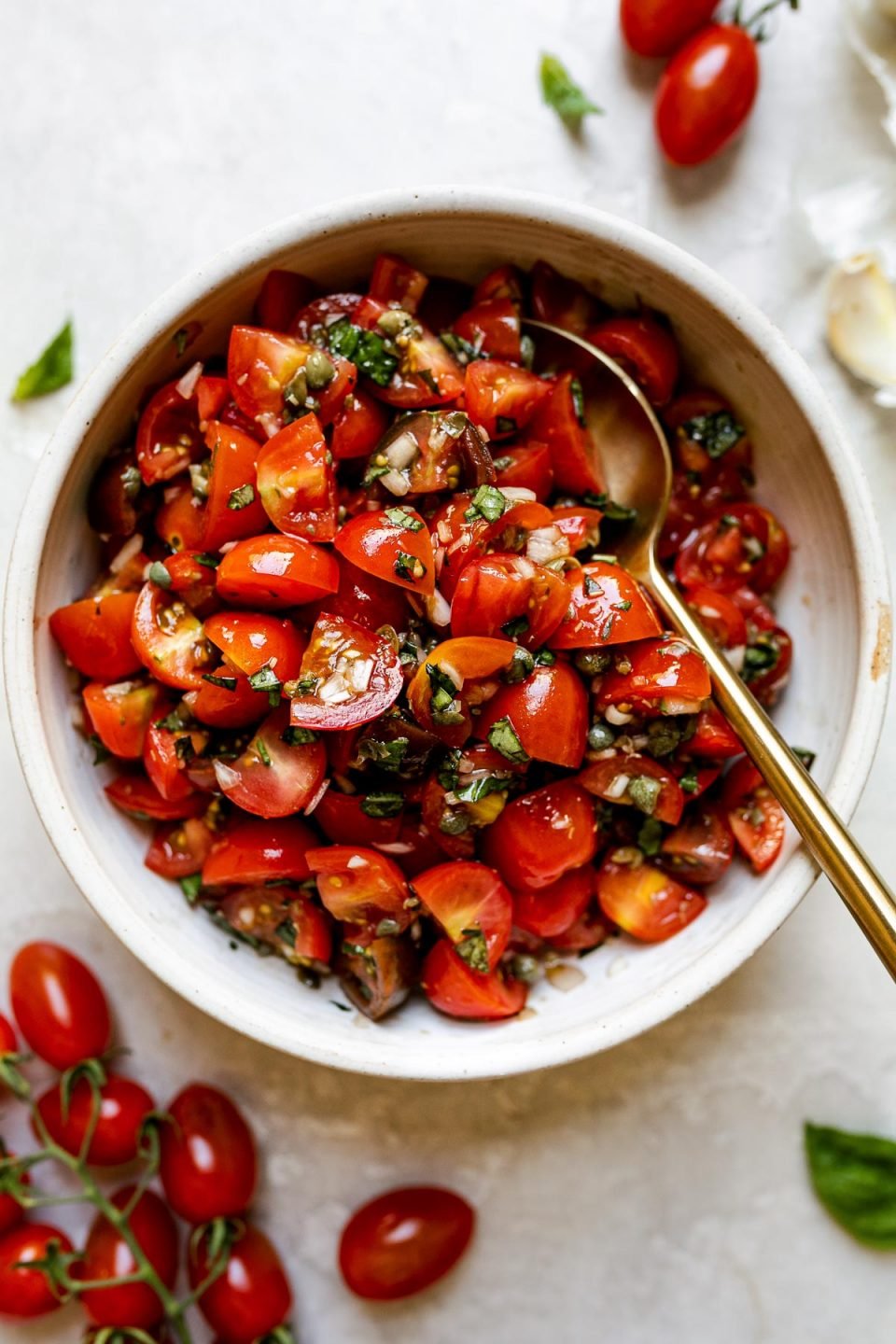 Top down view of fresh bruschetta in a small light colored ceramic bowl atop of a textured white surface. A gold spoon is nestled in the bowl, and the bowl is surrounded by fresh loose basil, cherry tomatoes on the vine, a few loose cherry tomatoes, & a clove of garlic.