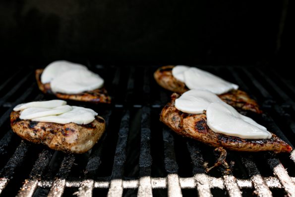 4 balsamic-marinated grilled chicken breasts being cooked on a gas grill. All four rest on the gas grill grates after being flipped once & have been topped with fresh mozzarella cheese that is beginning to melt over top.