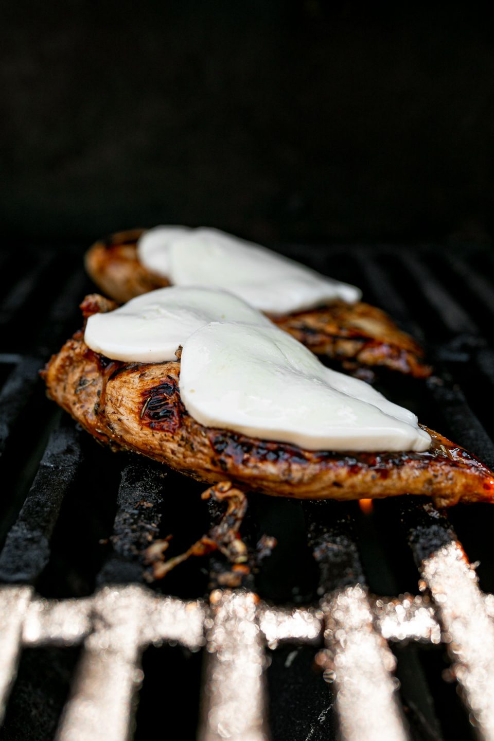 A close up of 2 balsamic-marinated grilled chicken breasts being cooked on a gas grill. The 2 chicken breasts rest on the gas grill grates after being flipped once & have been topped with fresh mozzarella cheese that is beginning to melt over top.