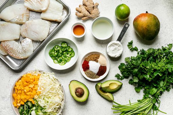 Grilled cod tacos ingredients arranged on a white surface – cod fillets, ginger, agave, lime, sour cream, mango, cilantro, avocado, spices, sliced jalapeno, shredded cabbage.