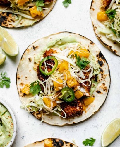 Assembled grilled cod fish tacos topped with ginger mango slaw. The tacos sit atop a white surface, surrounded by lime wedges, cilantro leaves, & a small dish of creamy avocado crema.