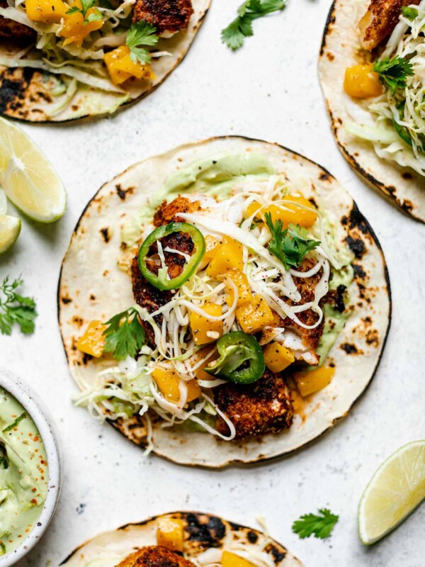 Assembled grilled cod fish tacos topped with ginger mango slaw. The tacos sit atop a white surface, surrounded by lime wedges, cilantro leaves, & a small dish of creamy avocado crema.