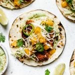 How to Make Grilled Cod Fish Tacos, Step 6: Serving. Assembled grilled cod fish tacos topped with ginger mango slaw. The tacos sit atop a white surface, surrounded by lime wedges, cilantro leaves, & a small dish of creamy avocado crema.