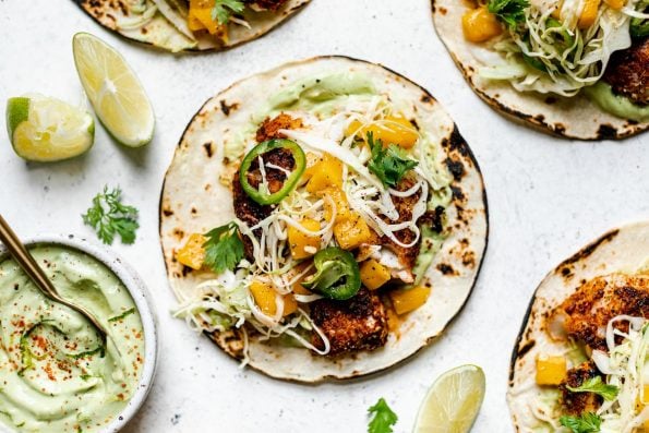 How to Make Grilled Cod Fish Tacos, Step 6: Serving. Assembled grilled cod fish tacos topped with ginger mango slaw. The tacos sit atop a white surface, surrounded by lime wedges, cilantro leaves, & a small dish of creamy avocado crema.