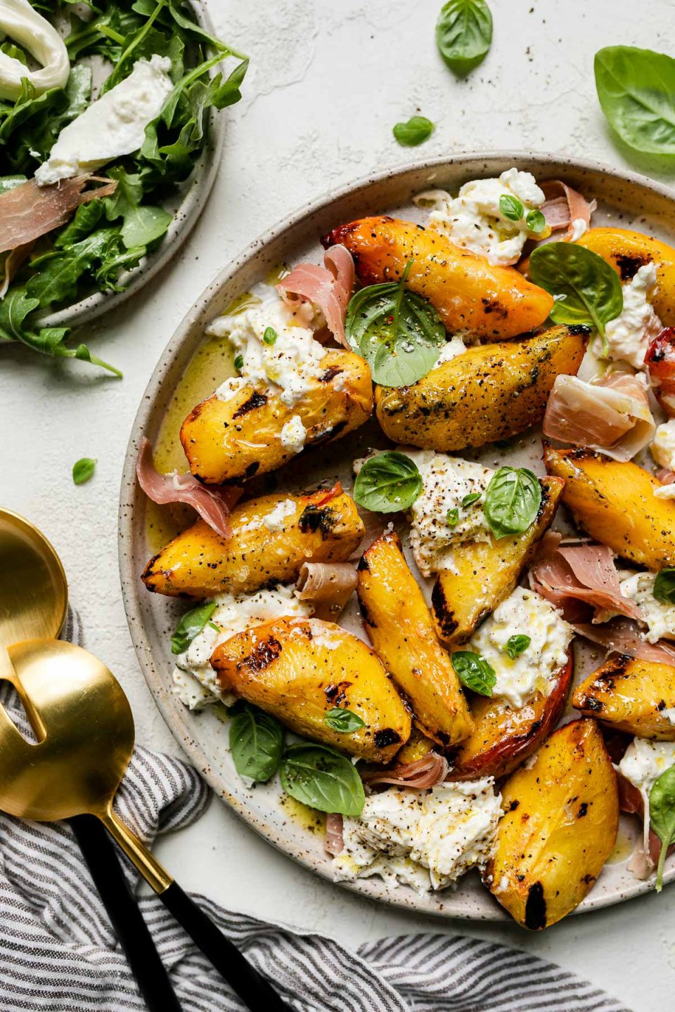 Grilled peaches, burrata cheese, fresh basil, & thinly sliced prosciutto arranged on a ceramic plate atop a textured white surface. Surrounding the grilled peach salad are fresh basil leaves, a small plate with arugula & grilled peach salad, a gray striped linen napkin, & large serving spoons.