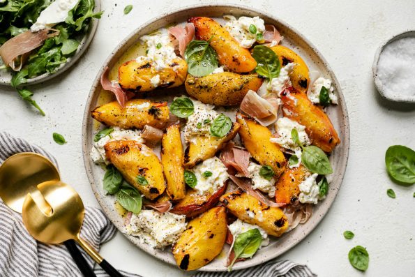 Grilled peaches, burrata cheese, fresh basil, & thinly sliced prosciutto arranged on a ceramic plate atop a textured white surface. Surrounding the grilled peach salad are fresh basil leaves, a small plate with arugula & grilled peach salad, a gray striped linen napkin, & large serving spoons.