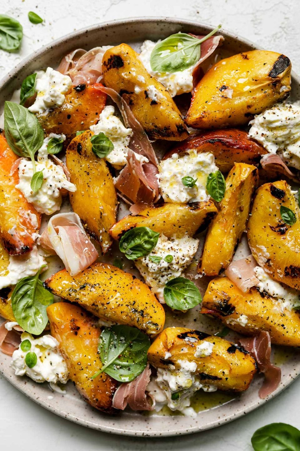 Grilled peaches, burrata cheese, fresh basil, & thinly sliced prosciutto arranged on a ceramic plate atop a textured white surface. Surrounding the grilled peach salad are fresh basil leaves.