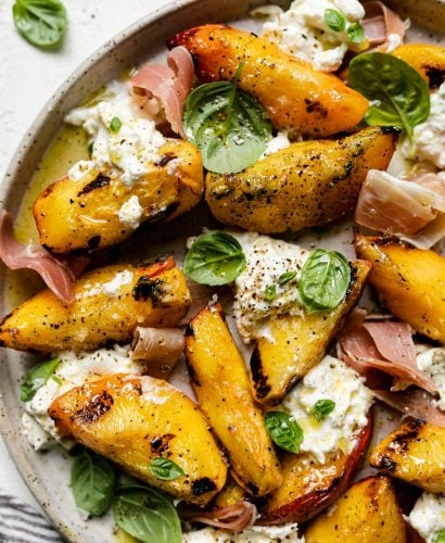 Grilled peaches, burrata cheese, fresh basil, & thinly sliced prosciutto arranged on a ceramic plate atop a textured white surface. Surrounding the grilled peach salad are fresh basil leaves & a gray striped linen napkin