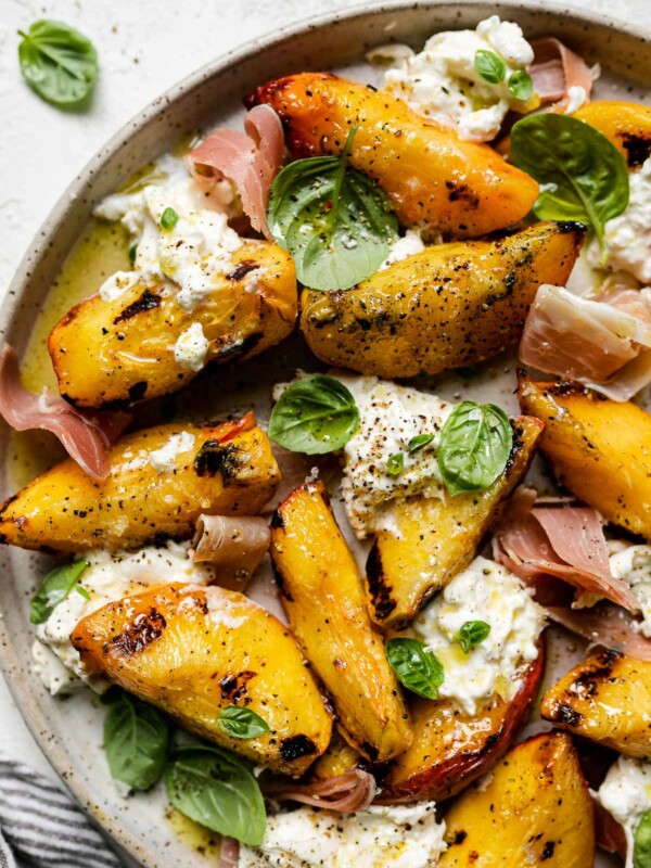 Grilled peaches, burrata cheese, fresh basil, & thinly sliced prosciutto arranged on a ceramic plate atop a textured white surface. Surrounding the grilled peach salad are fresh basil leaves & a gray striped linen napkin