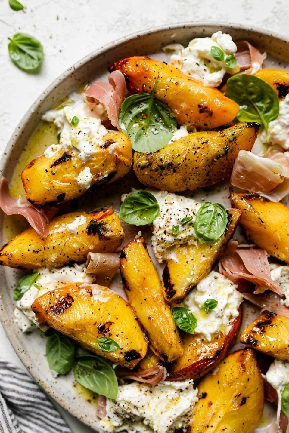 Grilled peaches, burrata cheese, fresh basil, & thinly sliced prosciutto arranged on a ceramic plate atop a textured white surface. Surrounding the grilled peach salad are fresh basil leaves & a gray striped linen napkin