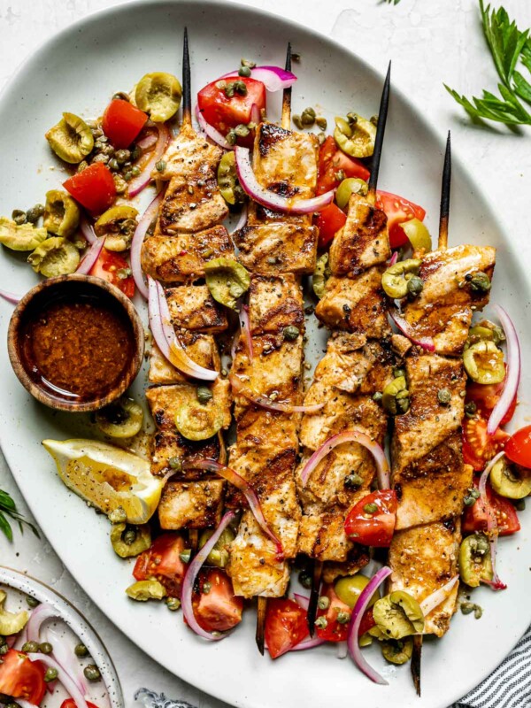 Grilled Swordfish Kabobs plated on a large serving platter with swordfish marinade as a serving sauce & a simple salad of olives, capers, tomatoes, & thinly sliced red onion. The platter sits atop a creamy cement surface alongside a striped gray linen napkin, a few sprigs of fresh parsley, & a small ceramic plate with the same pickley salad.