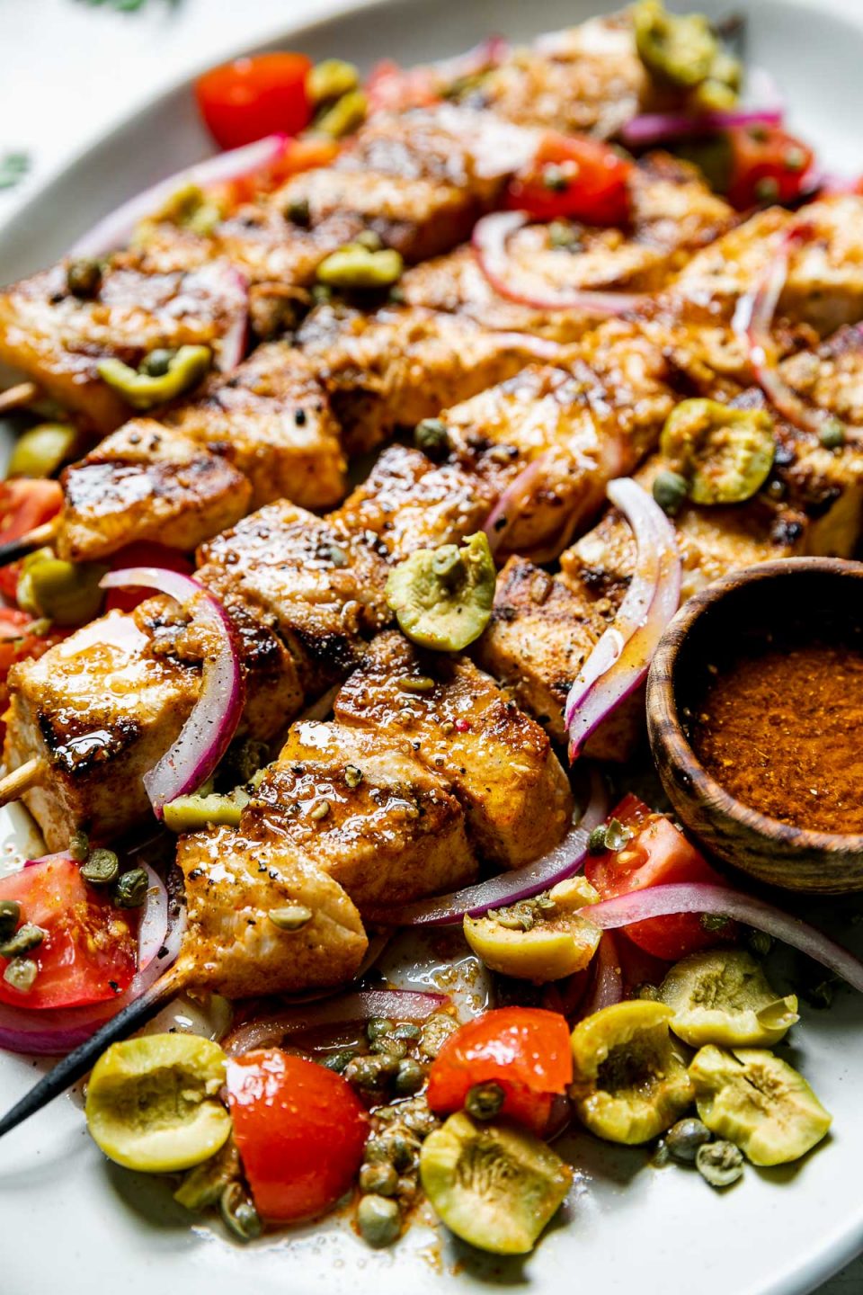 Grilled Swordfish Kabobs plated on a large serving platter with swordfish marinade as a serving sauce & a simple salad of olives, capers, tomatoes, & thinly sliced red onion.
