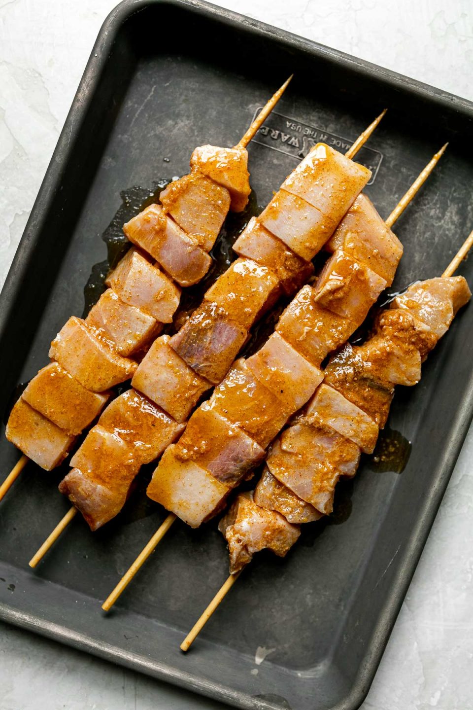Marinaded cubed swordfish threaded on 4 bamboo skewers sitting in a dark gray baking sheet atop a creamy cement surface.