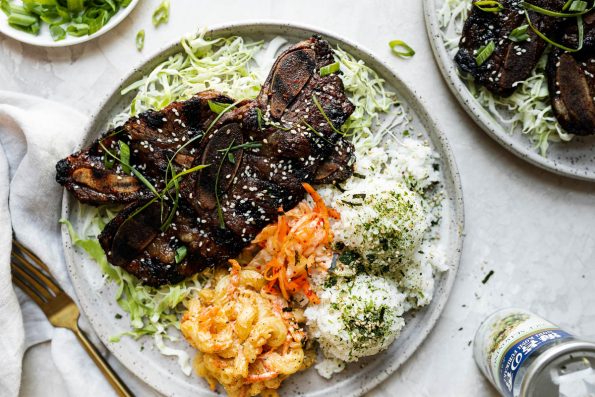 Kalbi served local Hawaiian-style, plated with shredded cabbage & scoops of rice, mac salad, & kimchi. The plate sits atop a cement surface alongside a second plate of kalbi, nori furikake seasoning, a gray linen napkin, & a plate of thin sliced green onions.