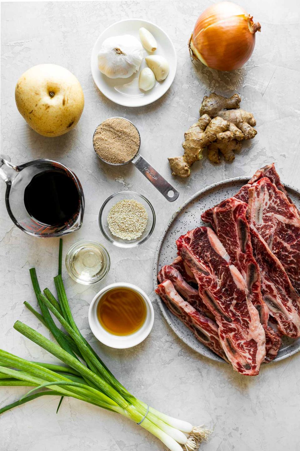 Grilled Kalbi ingredients arranged on a cement surface – green onions, sesame oil, rice vinegar, sesame seeds, garlic, brown sugar, onion, soy sauce, ginger, pear, & flanken-style beef short ribs.