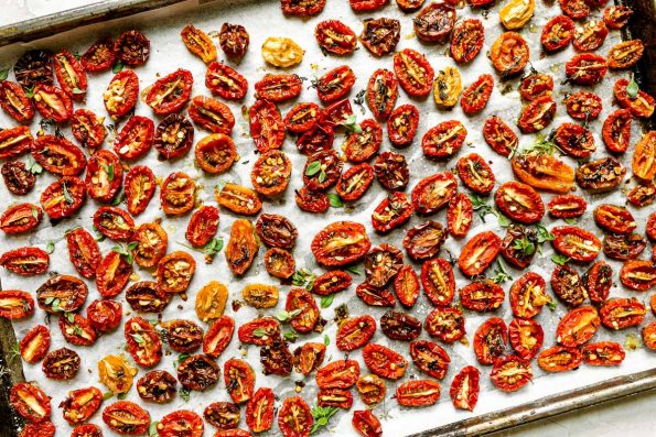 A close up of slow roasted cherry tomatoes arranged on a sheet pan lined with white parchment paper & sprinkled with fresh herbs.