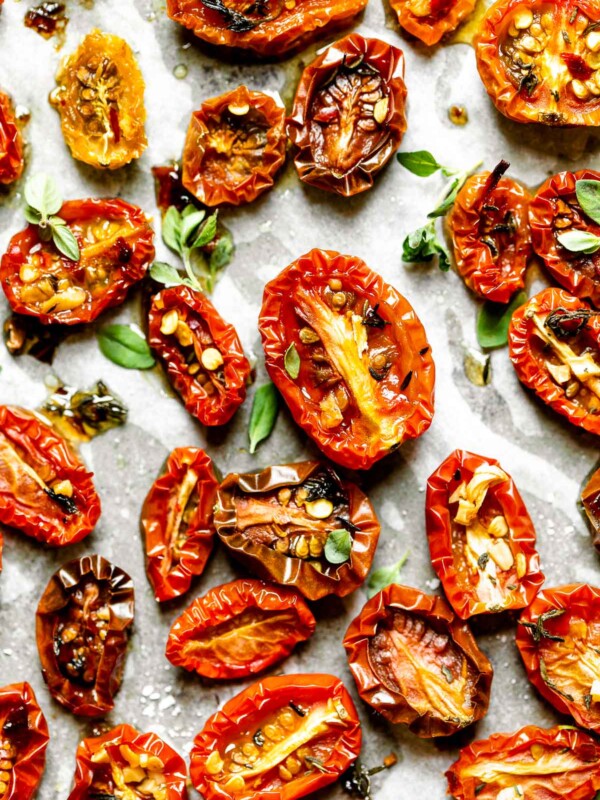 A close up of slow roasted cherry tomatoes arranged on a sheet pan lined with white parchment paper & sprinkled with fresh herbs.