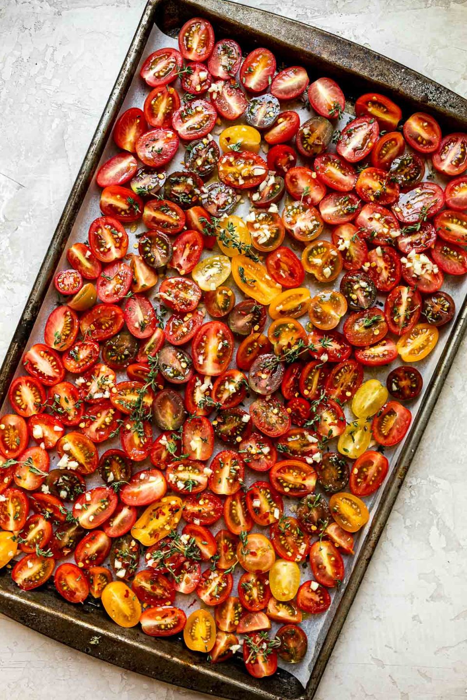 Raw cherry tomatoes arranged on a sheet pan lined with white parchment paper - sprinkled with fresh herbs & garlic ready to be slow roasted. The sheet pan rests on top of a light gray colored textured surface.