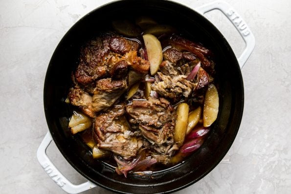 Apple Cider Braised Pork Shoulder, with softened apples & red onions, in a white Dutch oven atop a creamy cement surface.
