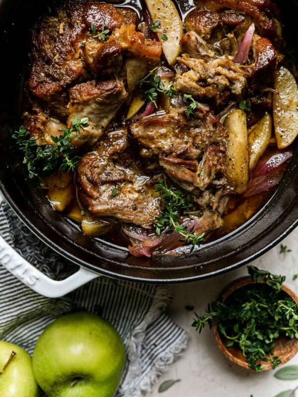 Apple Cider Braised Pork Shoulder in a white Dutch oven atop a creamy cement surface. Fresh thyme has been sprinkled on top. A small pinch bowl of additional fresh herbs, a white & blue striped linen napkin, & two granny smith apples sit alongside the Dutch oven.