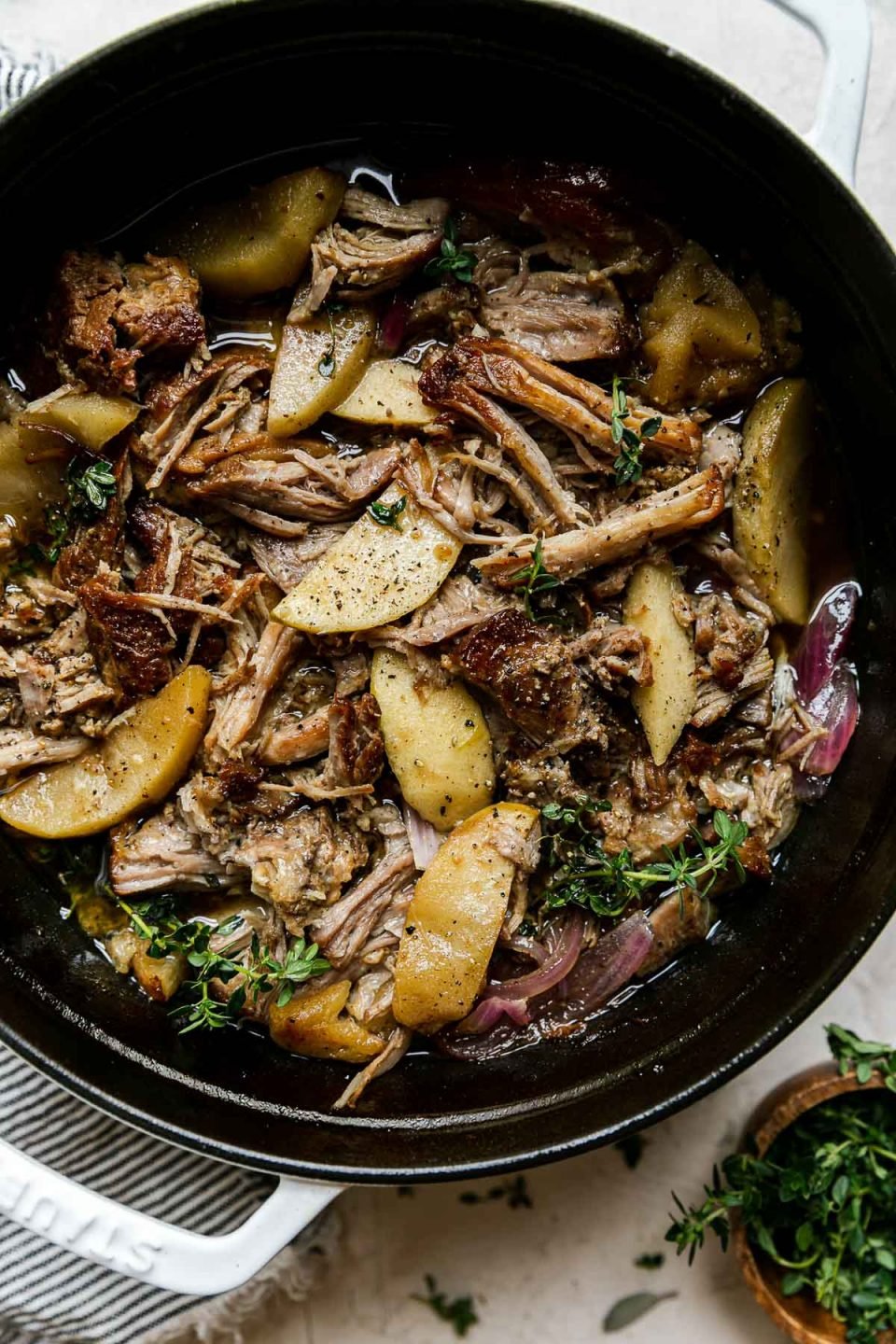 Apple Cider Braised Pork Shoulder in a white Dutch oven atop a creamy cement surface. Fresh thyme has been sprinkled on top. A small pinch bowl of additional fresh herbs, a white & blue striped linen napkin sit alongside the Dutch oven.
