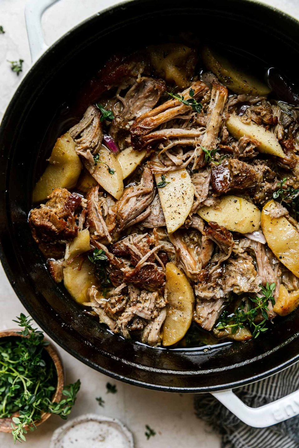 Apple Cider Braised Pork Shoulder in a white Dutch oven atop a creamy cement surface. Fresh thyme has been sprinkled on top. Two small pinch bowls filled with additional fresh herbs & kosher salt, & a white & blue striped linen napkin sit alongside the Dutch oven.