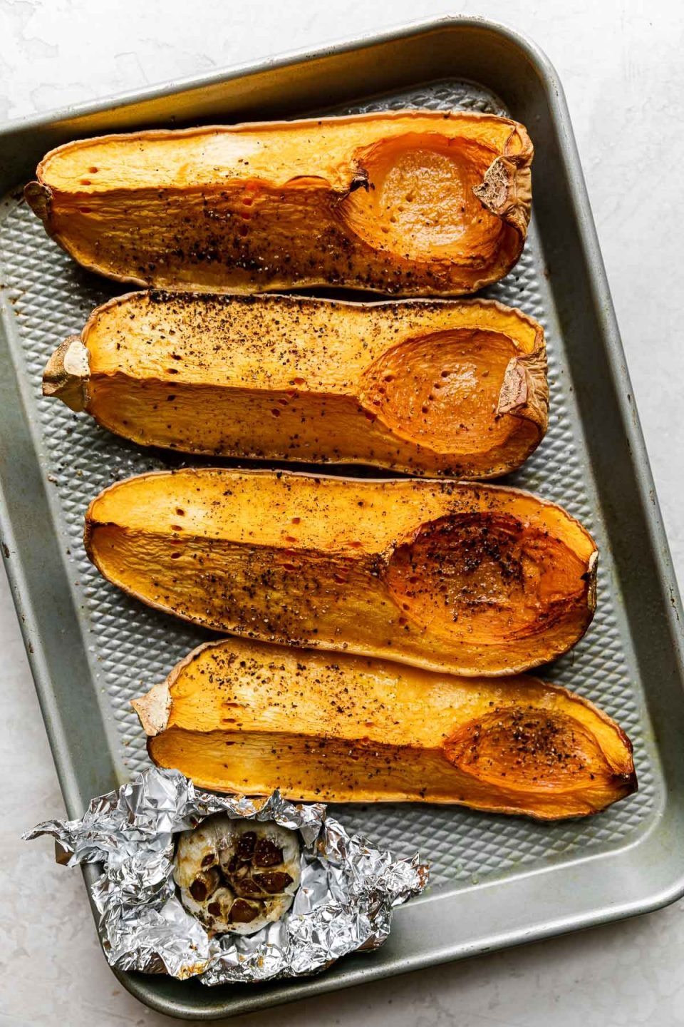 Four quartered & roasted butternut squash pieces arranged on a sheet pan alongside a bulb of roasted garlic wrapped in tin foil. The sheet pan rests on a creamy cement surface.