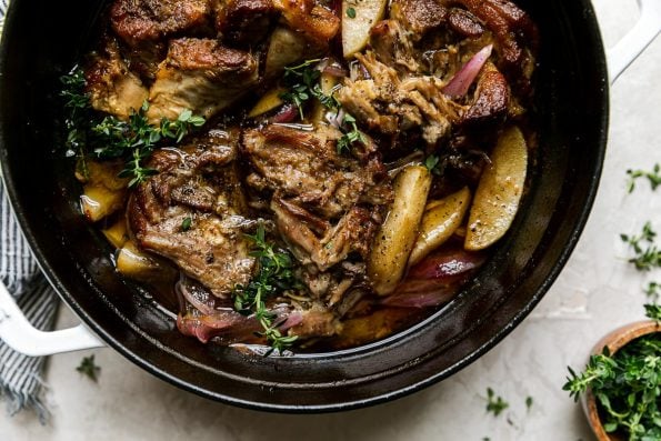 Apple Cider Braised Pork Shoulder in a white Dutch oven atop a creamy cement surface. Fresh thyme has been sprinkled on top and a few pieces rest on the cement surface. A white & blue striped linen napkin & a wooden pinch bowl filled with fresh herbs sits alongside.