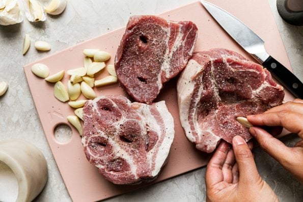 How to make Apple Cider Braised Pork Shoulder, step 1: a woman's hands shown studding pork shoulder roast with garlic cloves. the pork shoulder sits atop a light pink cutting board next to a pile of peeled garlic cloves & a container or kosher salt on a creamy cement surface.