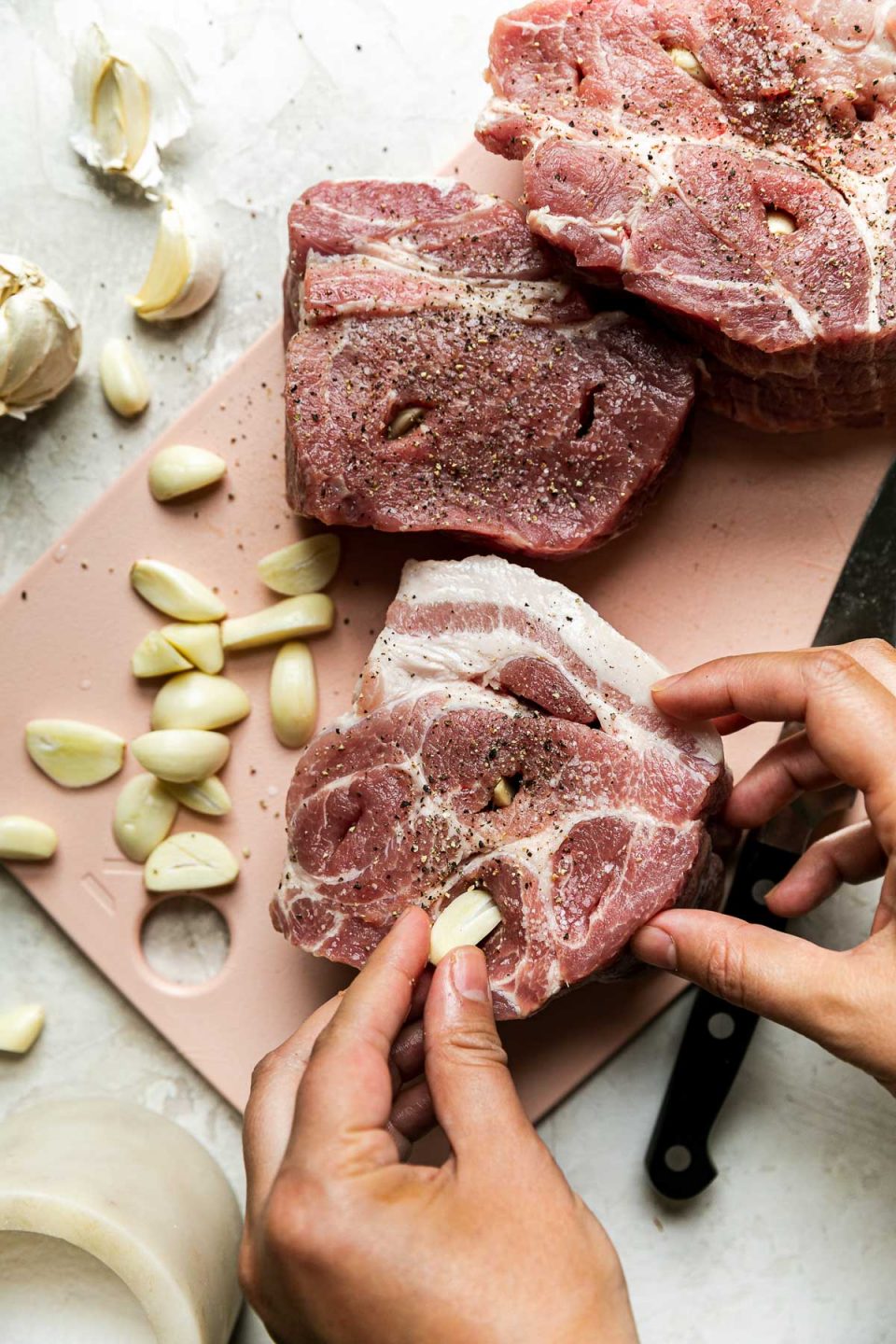 How to make Apple Cider Braised Pork Shoulder, step 1: a woman's hands shown studding pork shoulder roast with garlic cloves. the pork shoulder sits atop a light pink cutting board next to a pile of peeled garlic cloves & a container or kosher salt on a creamy cement surface.