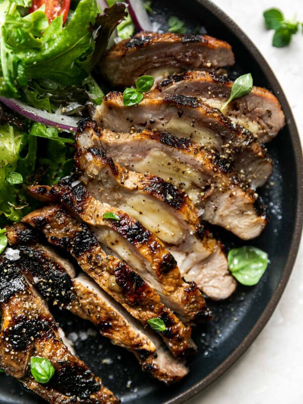 Grilled stuffed pork chop on a black plate. The pork chop is sliced, revealing its cheese & prosciutto filling. It's plated with a fresh green salad with tomatoes & red onions. The plate sits atop a creamy cement surface with a few sprigs of fresh oregano & a striped linen napkin.