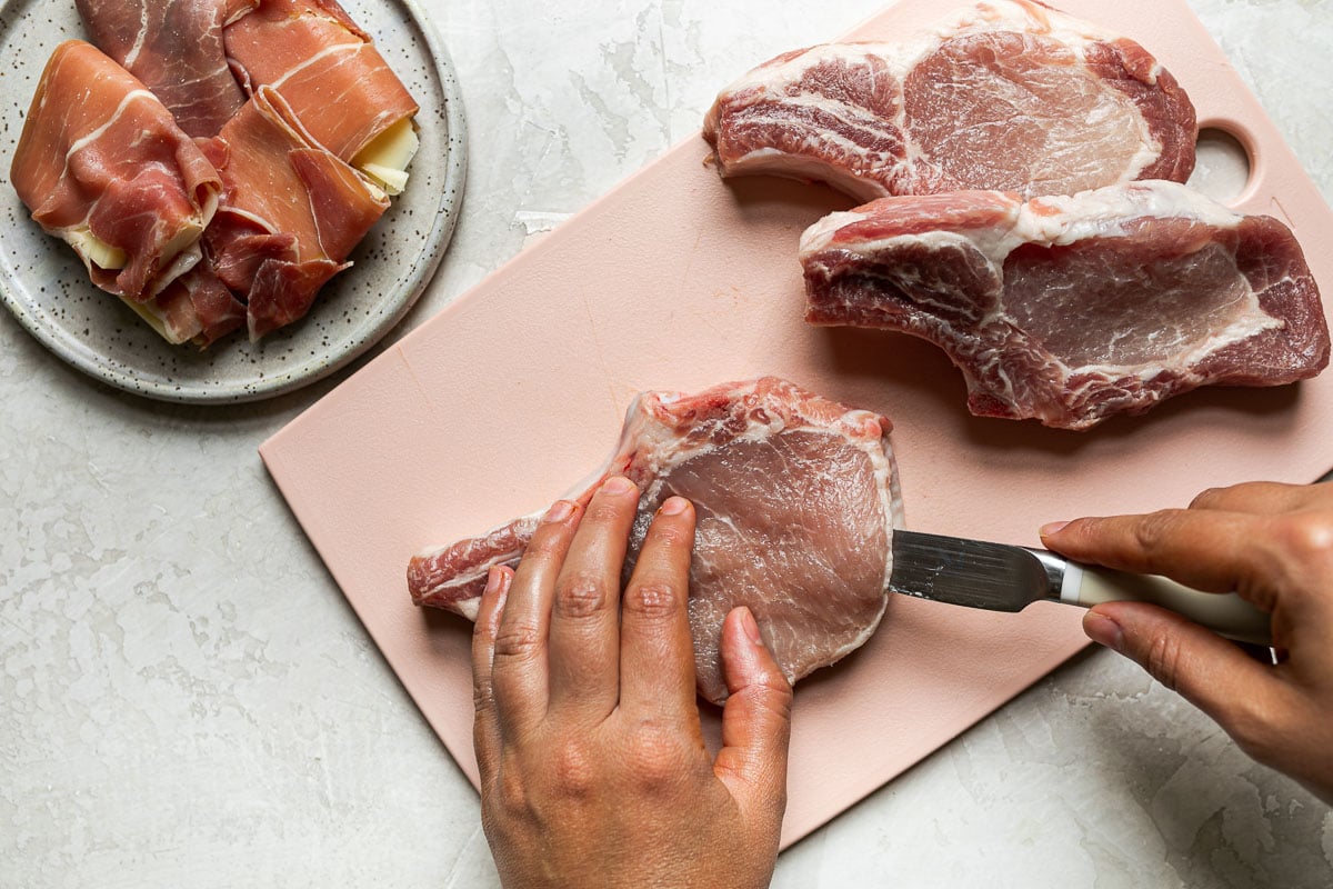 How to Stuff Bone-In Pork Chops – 3 bone-in pork chops on a pink cutting board on a creamy cement surface. A woman's hands hold one of the pork chops, using a paring knife to cut a pocket through its center. Next to the cutting board is a small plate of prosciutto-warapped asiago cheese.