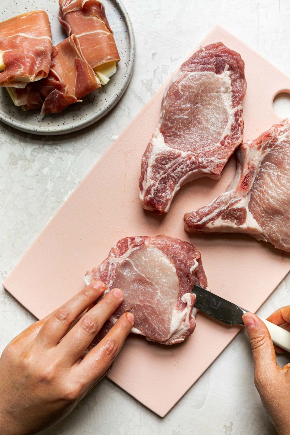 How to Stuff Bone-In Pork Chops – 3 bone-in pork chops on a pink cutting board on a creamy cement surface. A woman's hands hold one of the pork chops, using a paring knife to cut a pocket through its center. Next to the cutting board is a small plate of prosciutto-warapped asiago cheese.