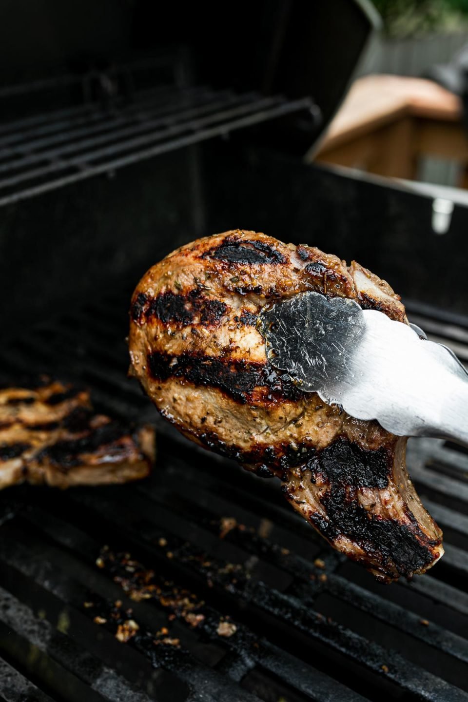 Grill tongs hold a grilled pork chop over grill grates.