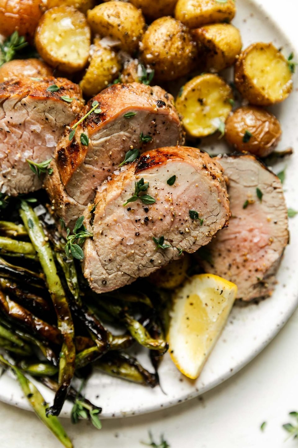 Lemon Garlic Grilled Pork Tenderloin on a white speckled ceramic plate. The tenderloin is sliced in medallions and garnished with fresh herbs. It's plated with baby golden potatoes, char-grilled green beans, & a lemon wedge. The plate sits atop a creamy cement surface.
