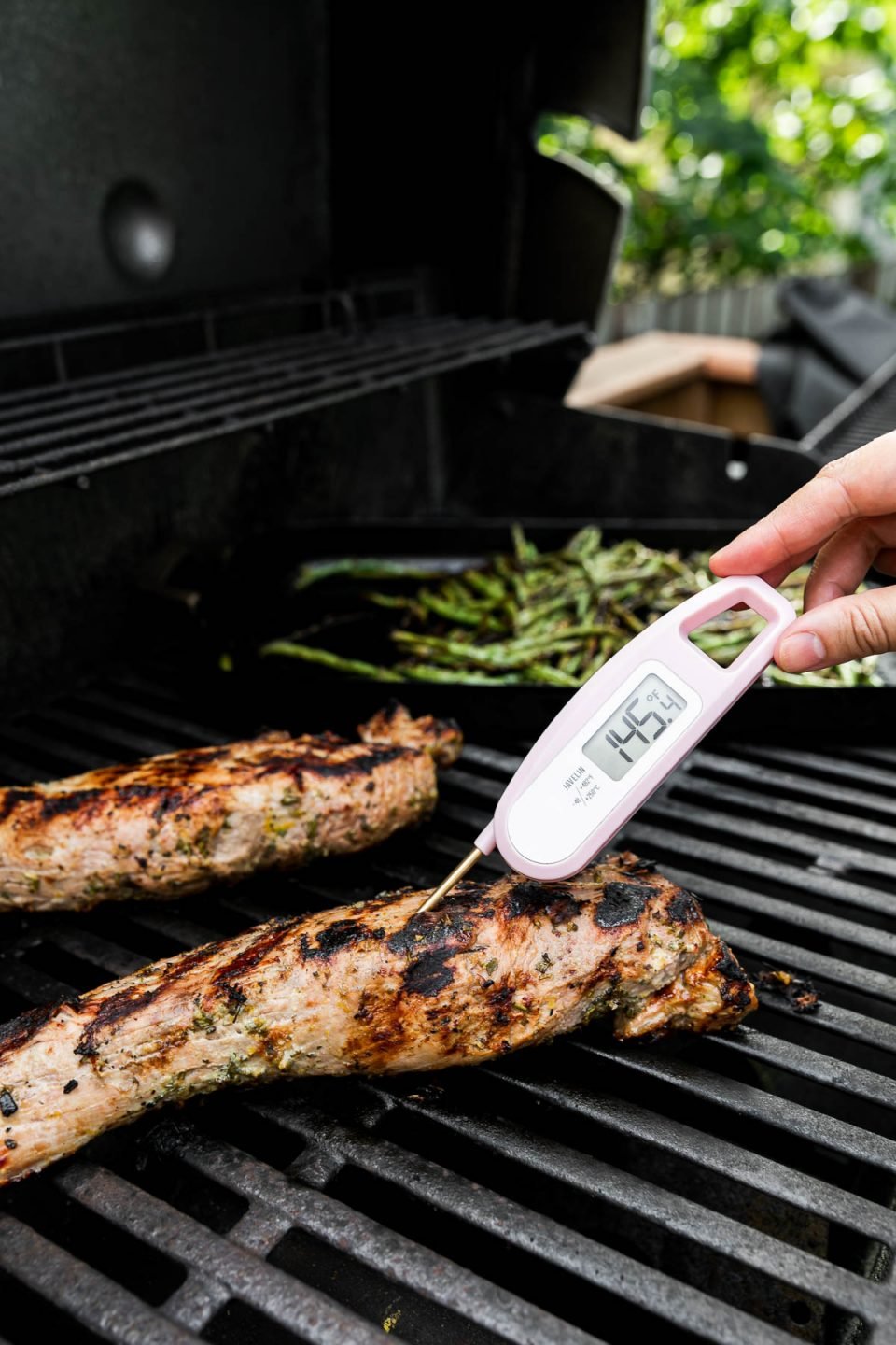 Two Lemon Garlic Grilled Pork Tenderloins on a gas grill. A woman's hand holds a pink instant read meat thermometer that is taking the internal temperature of one of the tenderloins. The thermometer registers at 145 degrees F. A grill basket rests on the grill grates in the background with char-grilled green beans cooking.