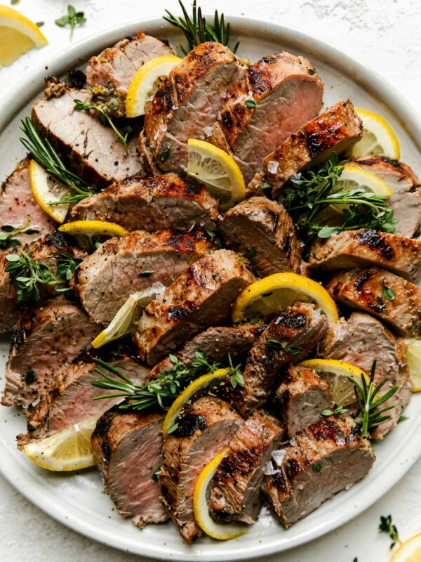 Lemon Garlic Grilled Pork Tenderloin on a white speckled ceramic plate. The tenderloin is sliced in medallions and garnished with sprigs of fresh rosemary, thyme, & lemon quarters for serving. The plate sits atop a creamy cement surface with lemon wedges & a few fresh herbs surrounding the plate & resting on the surface.