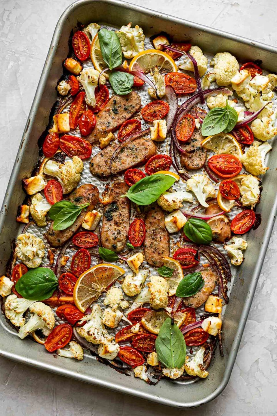 Baked Sheet Pan Chicken Sausage and Veggies on a silver quarter sheet pan atop a creamy cement surface. The sheet pan is topped with fresh basil leaves & ground black pepper.