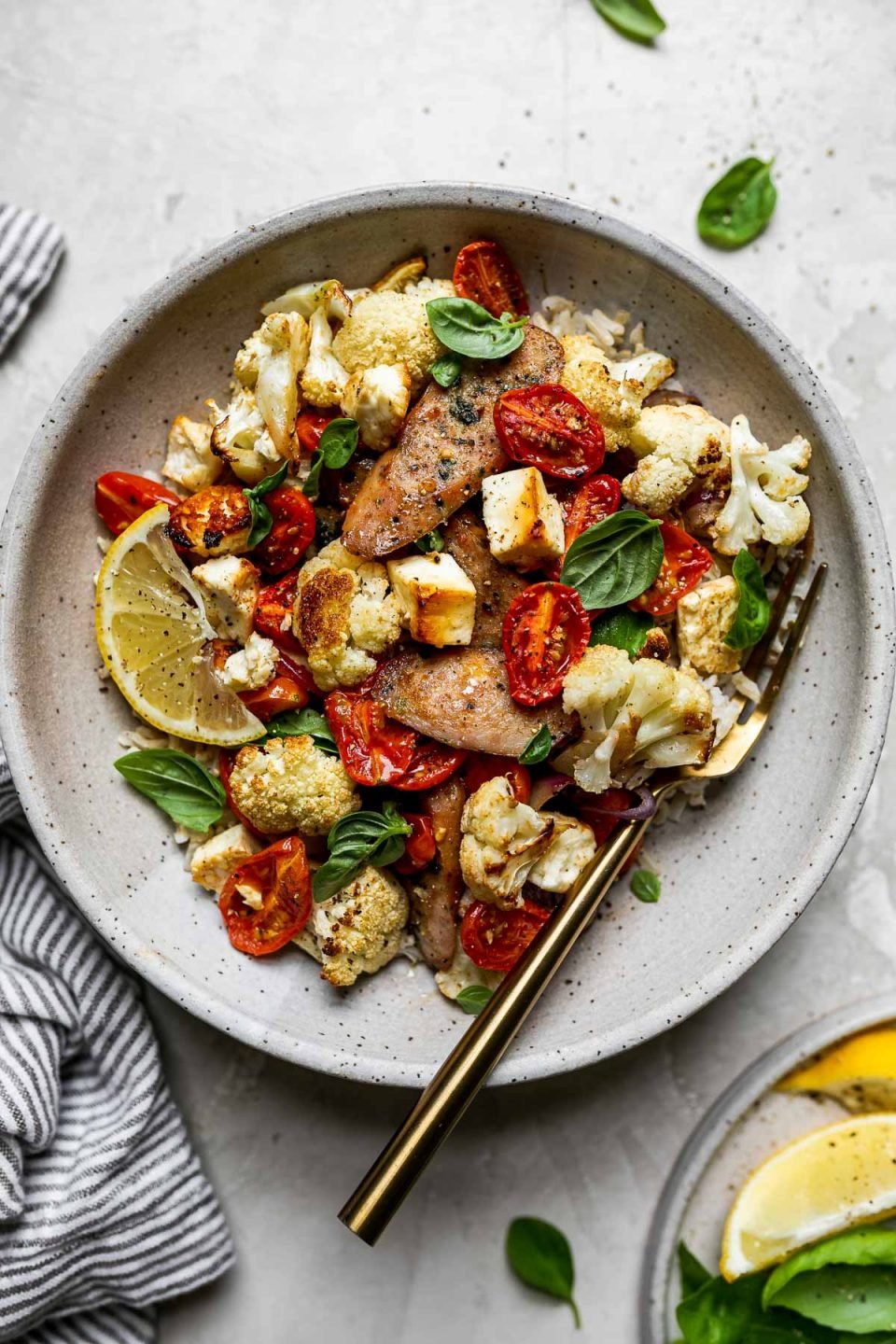 Sheet Pan Chicken Sausage and Veggies plated in a gray ceramic bowl over rice. The bowl has a gold fork in it and sits atop a creamy cement surface alongside a gray striped linen napkin, fresh basil leaves, & a small plate of lemon wedges & basil leaves for serving.