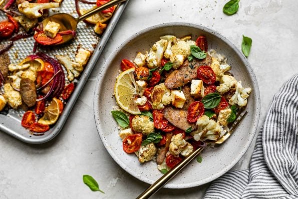 Sheet Pan Chicken Sausage and Veggies plated in a gray ceramic bowl over rice. The bowl has a gold fork in it and sits atop a creamy cement surface alongside a gray striped linen napkin, fresh basil leaves, & a small silver baking sheet with the baked chicken sausage & veggies.