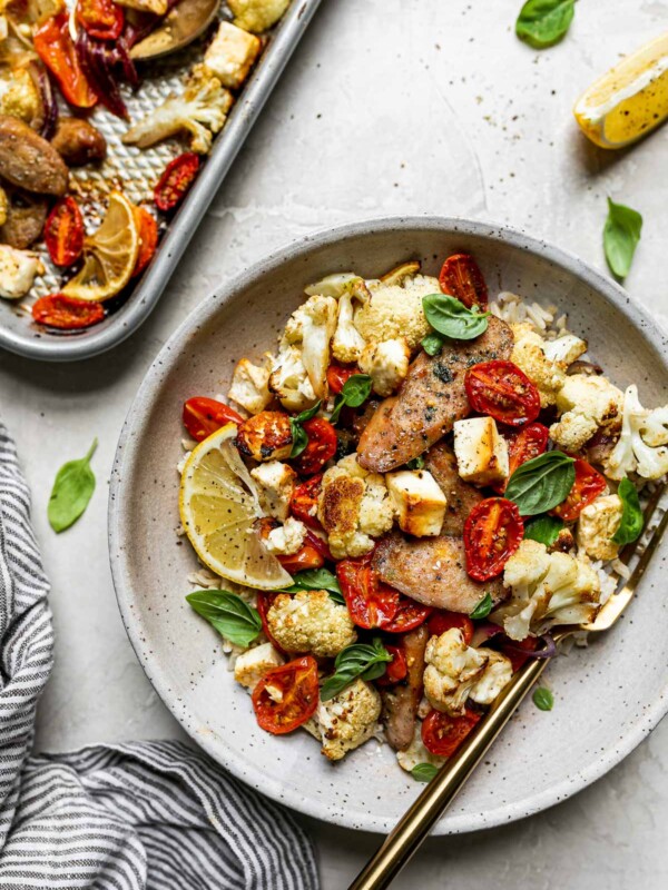 Sheet Pan Chicken Sausage and Veggies plated in a gray ceramic bowl over rice. The bowl has a gold fork in it and sits atop a creamy cement surface alongside a gray striped linen napkin, fresh basil leaves, & a small silver baking sheet with the baked chicken sausage & veggies.