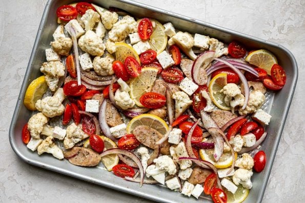 Sheet Pan Chicken Sausage and Veggies ingredients arranged on a silver quarter sheet pan atop a creamy cement surface.