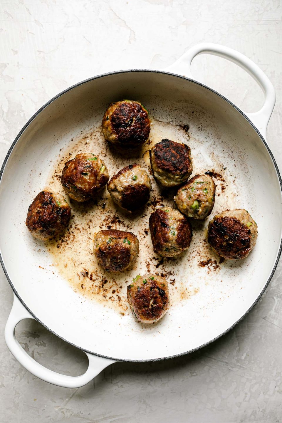 How to make ginger meatballs – browned meatballs in white enameled braising pan atop a creamy cement surface.